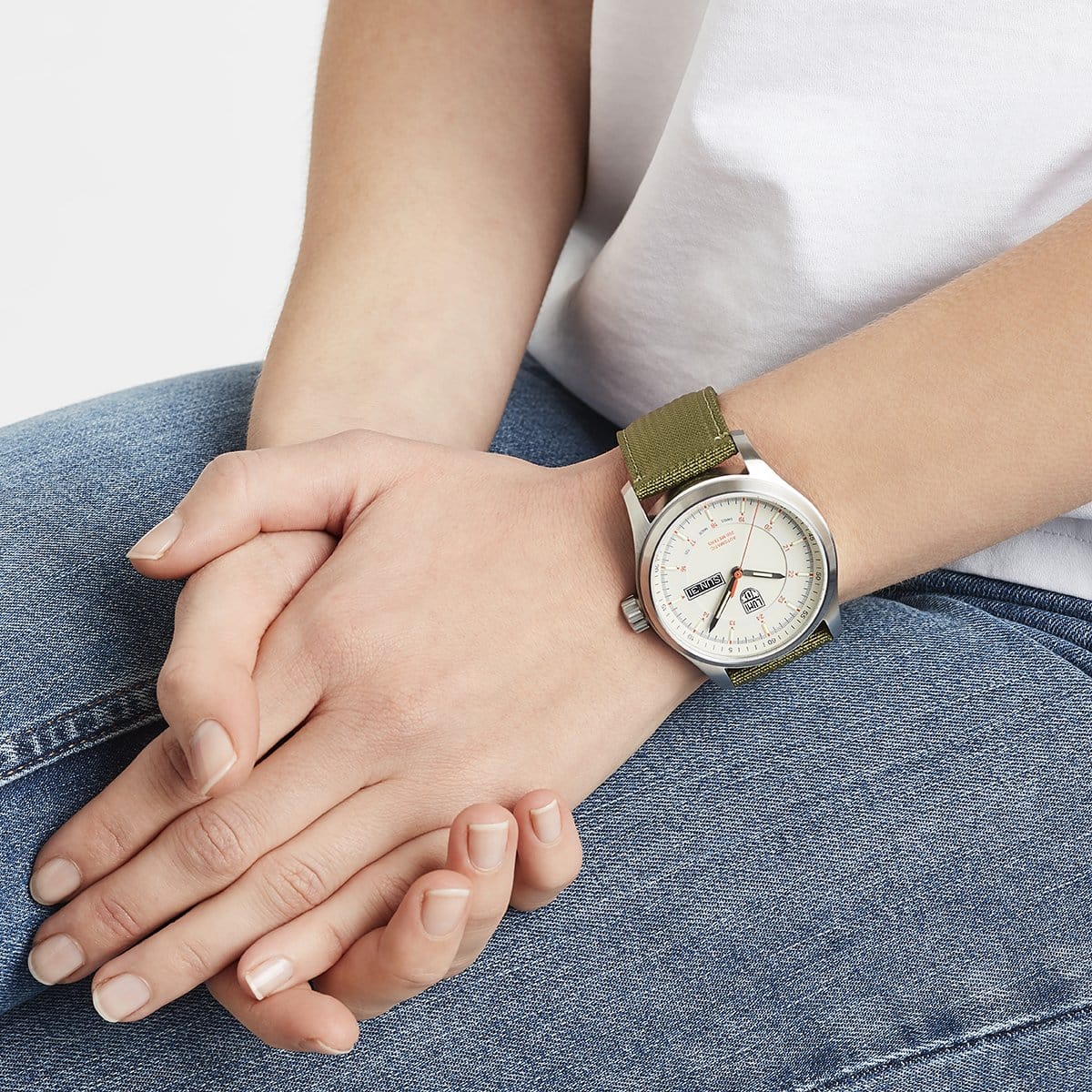 A person in blue jeans and a white shirt sits with folded hands, displaying a Luminox Atacama Field Automatic XL.1907.NF watch featuring a green fabric strap and light-colored dial.