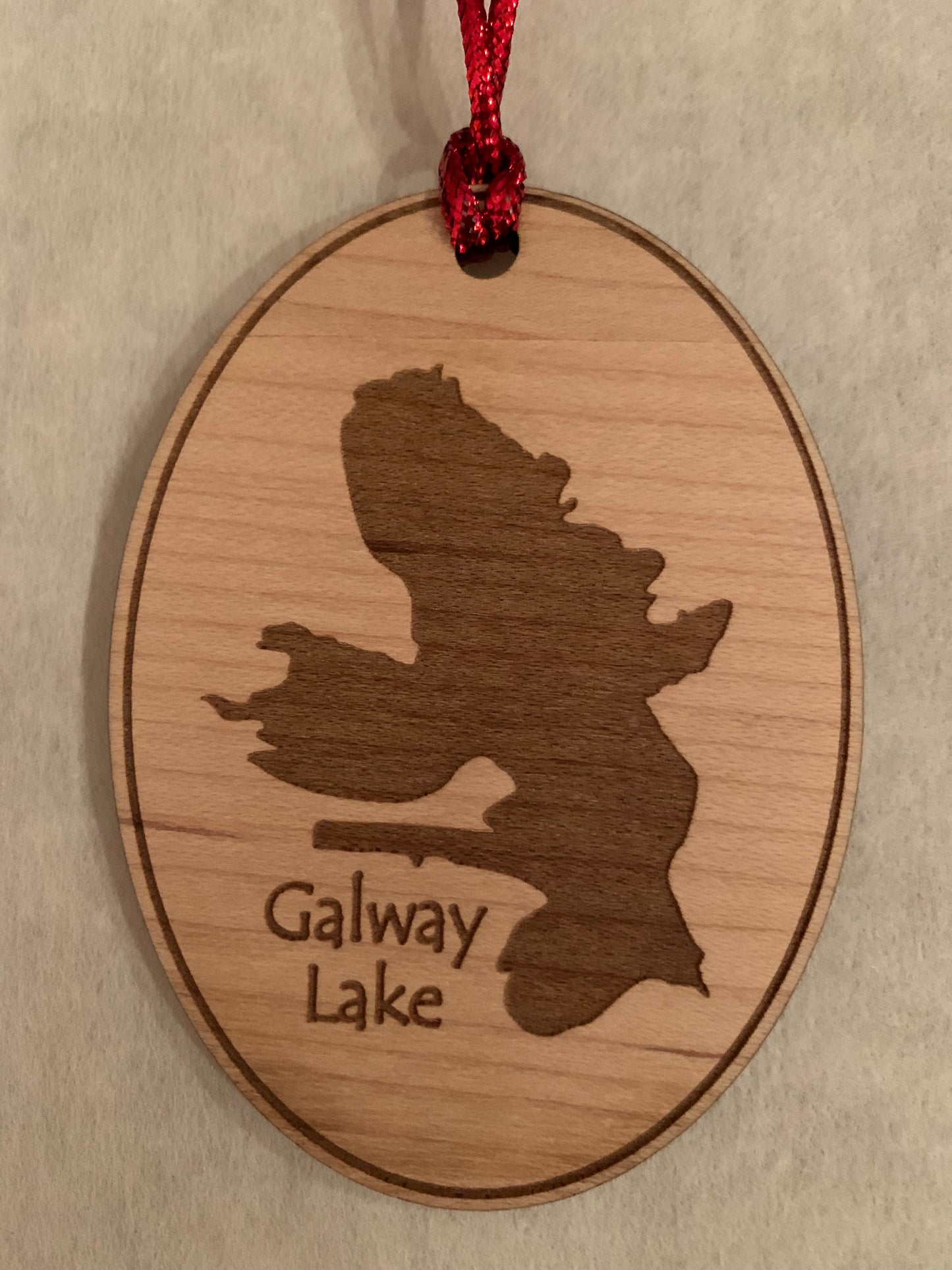 The Lake Art TS Ornament is a custom wooden Christmas ornament featuring an engraved outline of Galway Lake and the text "Galway Lake" below. It hangs from a red ribbon, making it a perfect keepsake for your tree.