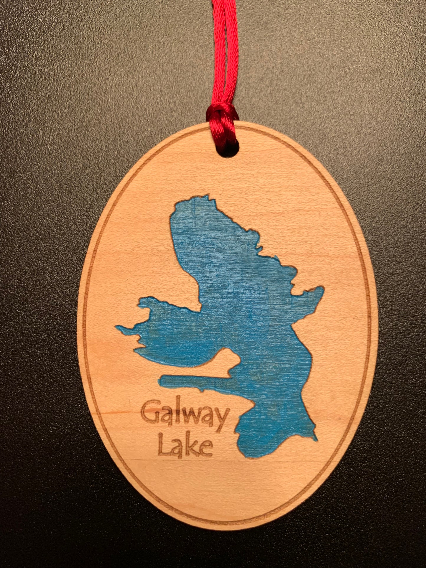 The Time Square Clock Shop’s TS Ornament - Custom Lake Christmas Ornament with Blue Water features Galway Lake’s silhouette and name, engraved on oval wood, hanging by a red ribbon on a black background.