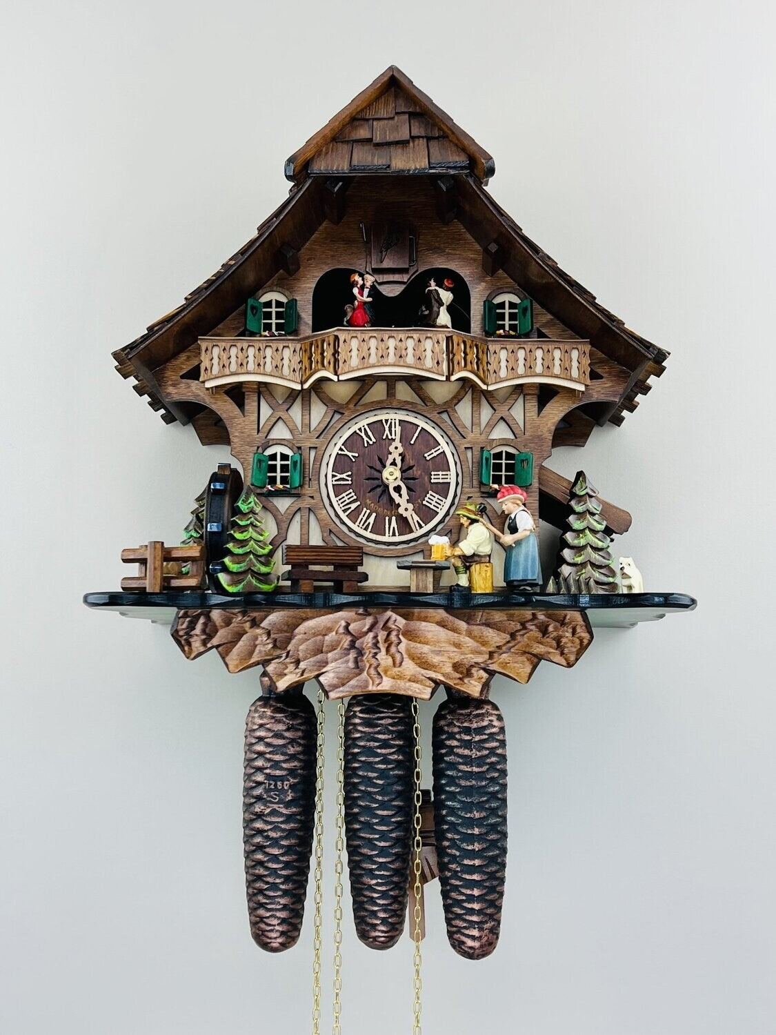 The River City Cuckoo Clock - The Noodle Bruiser (MD884-15) by River City Clocks features carved pine trees, a chalet-style house, balcony figurines, Roman numerals, pinecone weights, and chains set against a light background.