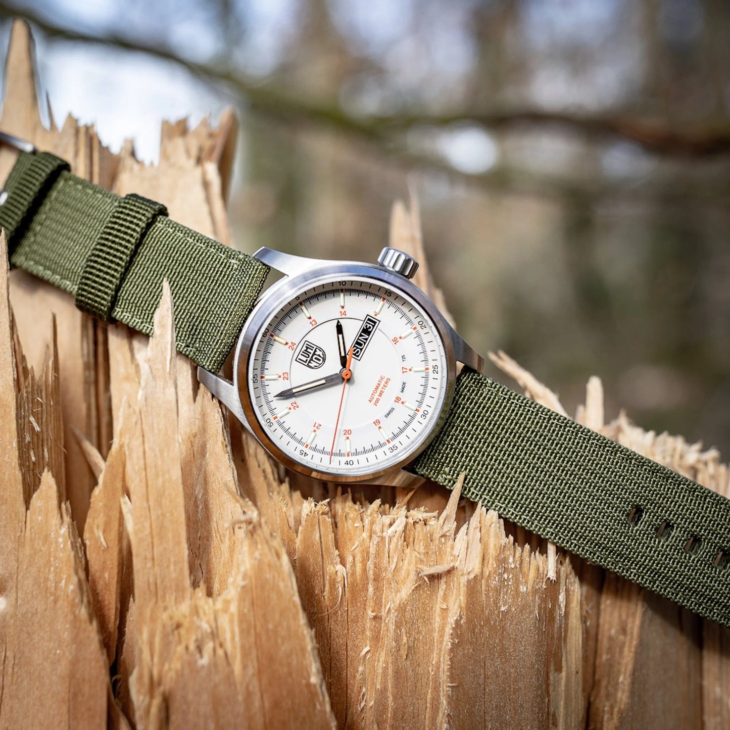 The Luminox Atacama Field Automatic XL.1907.NF, a CO₂ neutral watch with a white dial, orange accents, and green fabric strap, is displayed on jagged wood outdoors against a softly blurred natural background.