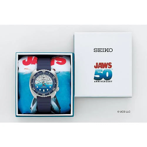 The Seiko Prospex JAWS 50th Anniversary Limited Edition SRPL81 diver’s watch features a blue strap, ocean-inspired design, and comes in a special gift box beside a lid marked “SEIKO” and “JAWS 50th ANNIVERSARY.”.