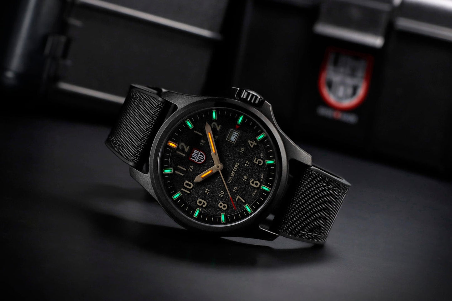 A Luminox Atacama Field 1970 (XL.1970) watch in black CARBONOX with a textured strap and glowing green and orange hour markers rests on a dark surface, blurred in the background is a red and white shield logo.