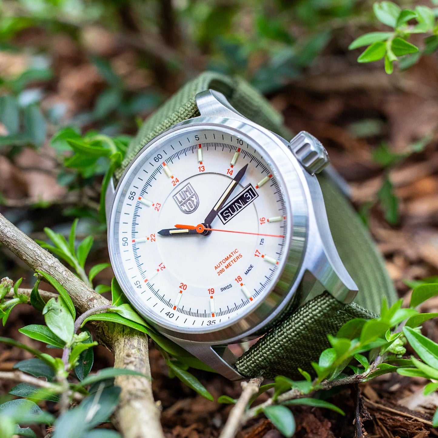 A Luminox Atacama Field Automatic XL.1907.NF silver watch with a white dial, orange second hand, green fabric strap, and day-date display is placed outdoors among green plants and brown mulch.