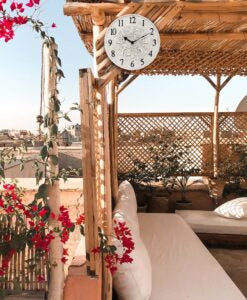 A cozy outdoor patio with bamboo furniture, white cushions, plants, red bougainvillea, and an Infinity Instruments Banc Fleur Clock (13377ST-C) showing 2:35 under a clear blue sky.