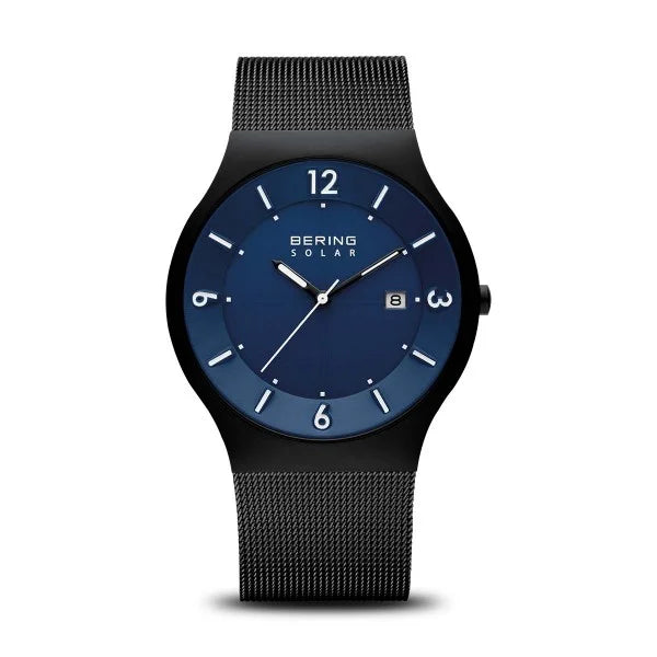 The Bering Watch - Slim Solar Collection 14440-227 features a blue dial, white hour markers, date display at 3 o’clock, and a black mesh band. Its round, sleek case adds to the modern minimalist style from Bering.