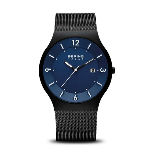 The Bering Watch - Slim Solar Collection 14440-227 features a blue dial, white hour markers, date display at 3 o’clock, and a black mesh band. Its round, sleek case adds to the modern minimalist style from Bering.