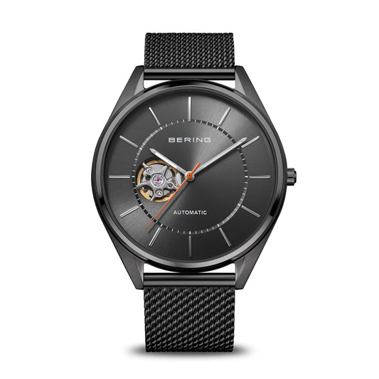 The Bering Watch - Automatic - 16743-377 by Bering features a sleek black stainless steel case, a mesh band, a minimalist dial with silver hour markers, an orange second hand, and an open heart design at 9 o’clock that reveals the watch’s automatic movement.