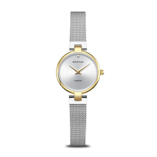 The Bering Watch - Classic Collection 17724-010 is a minimalist women's watch featuring a silver mesh band, round white dial, gold bezel, subtle markings, and a single diamond at 12 o’clock with the BERING logo near the top.