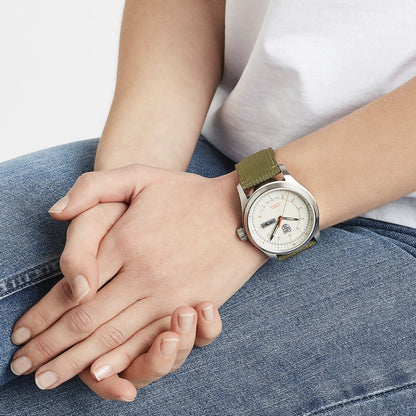 A person in blue jeans and a white shirt sits with folded hands, displaying a Luminox Atacama Field Automatic XL.1907.NF watch featuring a green fabric strap and light-colored dial.
