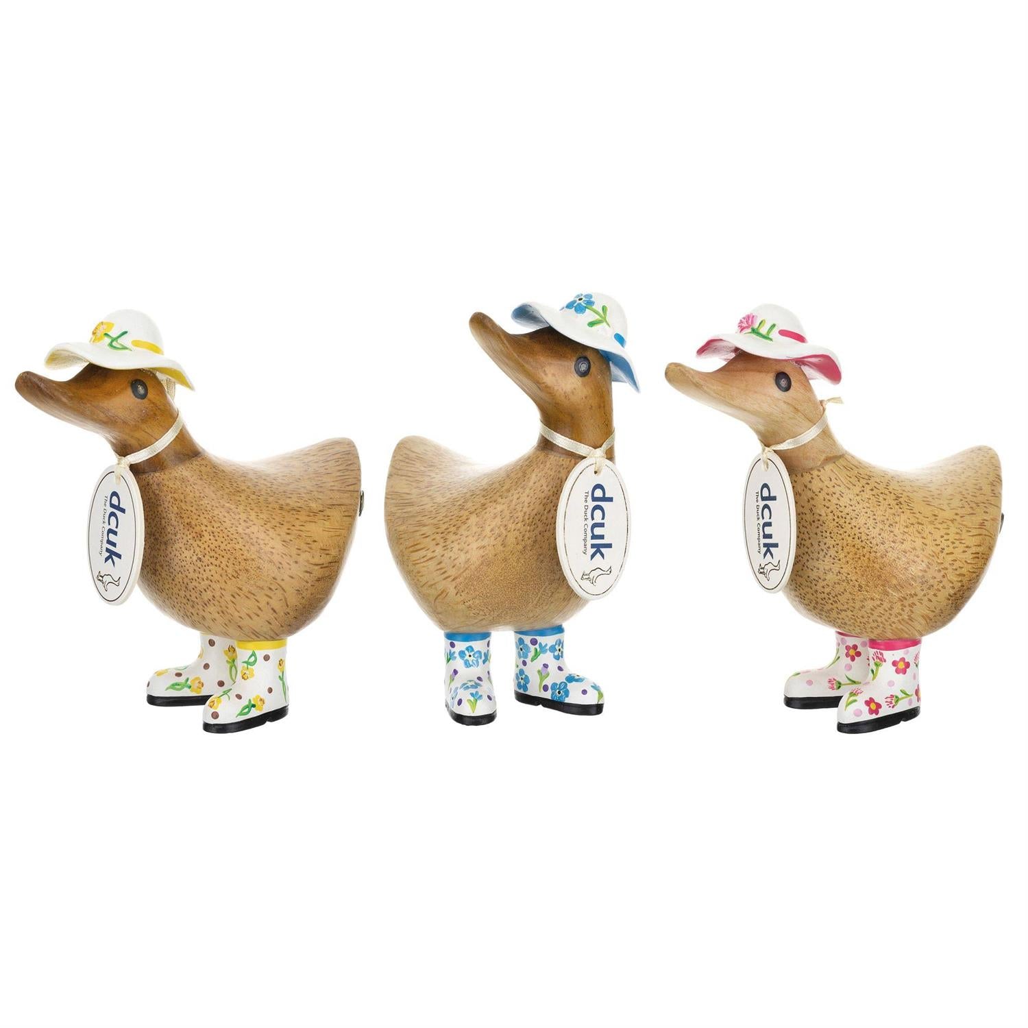 Three Enesco DCUK Ducks—Duckys with white hats and floral boots (6019178)—stand in a row, each charming bamboo duckling featuring unique accessory colors and a DCUK tag around its neck.