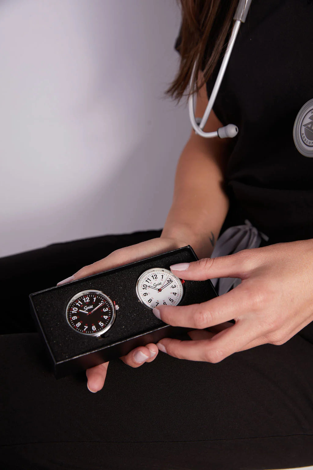 A person with a stethoscope holds a black box containing the Speidel Watch - Scrub FOB Watch set of 2 (model 60341000), featuring analog clip-on nurse watches with one black face and one white face, by Speidel.