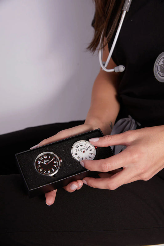 A person with a stethoscope holds a black box containing the Speidel Watch - Scrub FOB Watch set of 2 (model 60341000), featuring analog clip-on nurse watches with one black face and one white face, by Speidel.