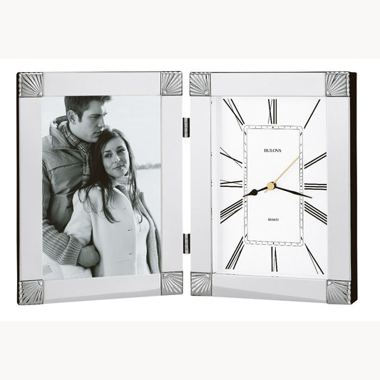 A framed photo of a couple in winter clothing stands beside the elegant Bulova Ceremonial Clock (B1254), both set in a hinged chrome finish display with Roman numerals and a sleek engraving plate.