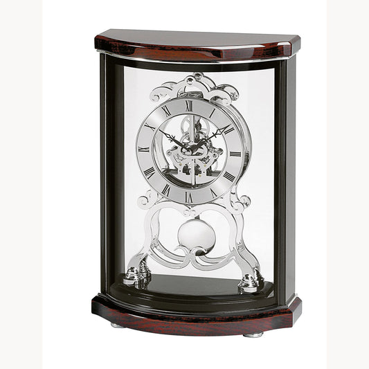 The Bulova Wentworth B2025 clock features a glass case, exposed silver skeleton movement, black Roman numerals, and visible pendulum. Its wooden frame has a dark mahogany piano finish with curved edges for added elegance.