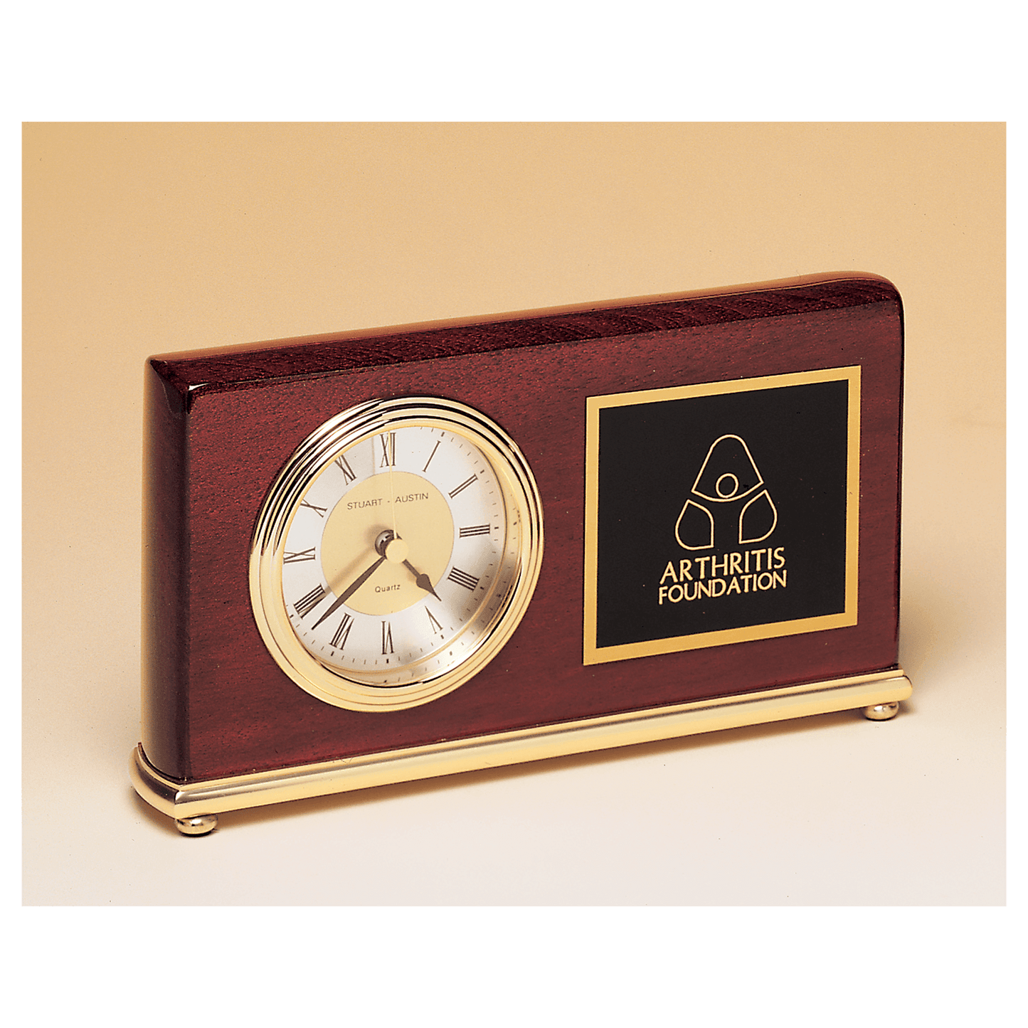 The JDS Desk Clock - Rosewood Piano Finish Clock (BC48) features a round gold-trimmed face, brass base, and an Arthritis Foundation logo plaque on the right, accented with elegant gold details.