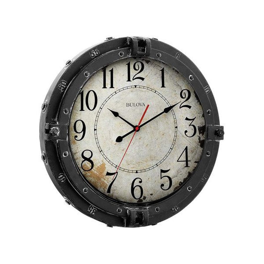 The Bulova Clock - Navigator (C4823) by Bulova features a weathered finish, distressed white face, black numbers and hands, and a dark metal frame with bolt details, reminiscent of a maritime porthole clock.