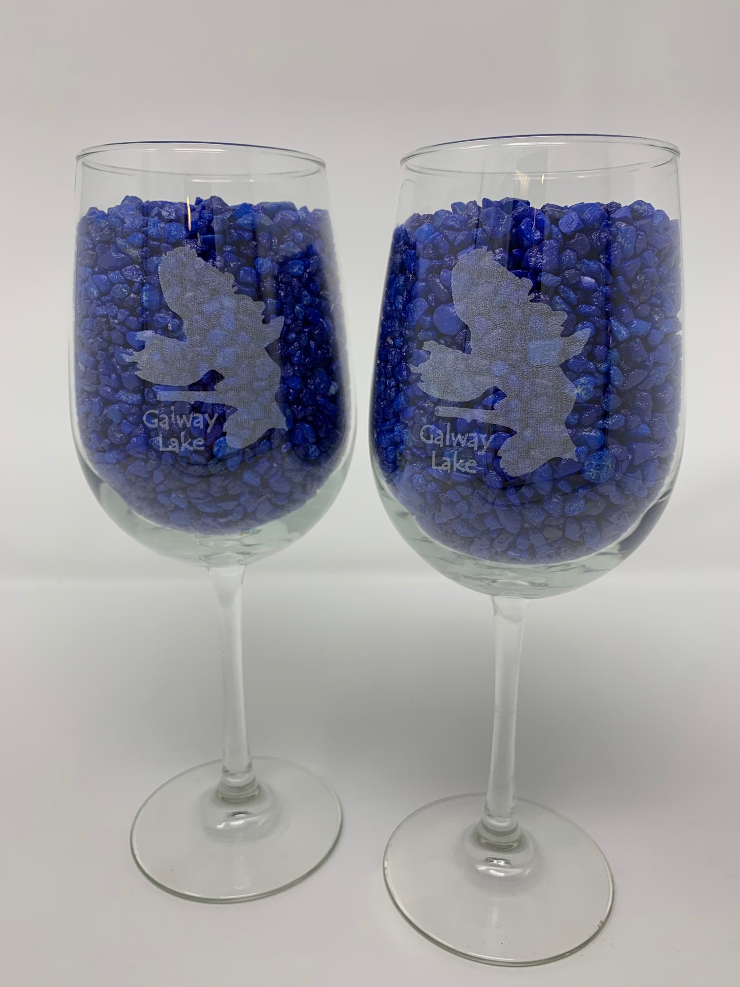 The Time Square Clock Shop TS Wine Glasses - Custom Set of 2 features each wine glass etched with a leaping person and "Galway Lake," filled with small blue stones, shown against a plain white background.