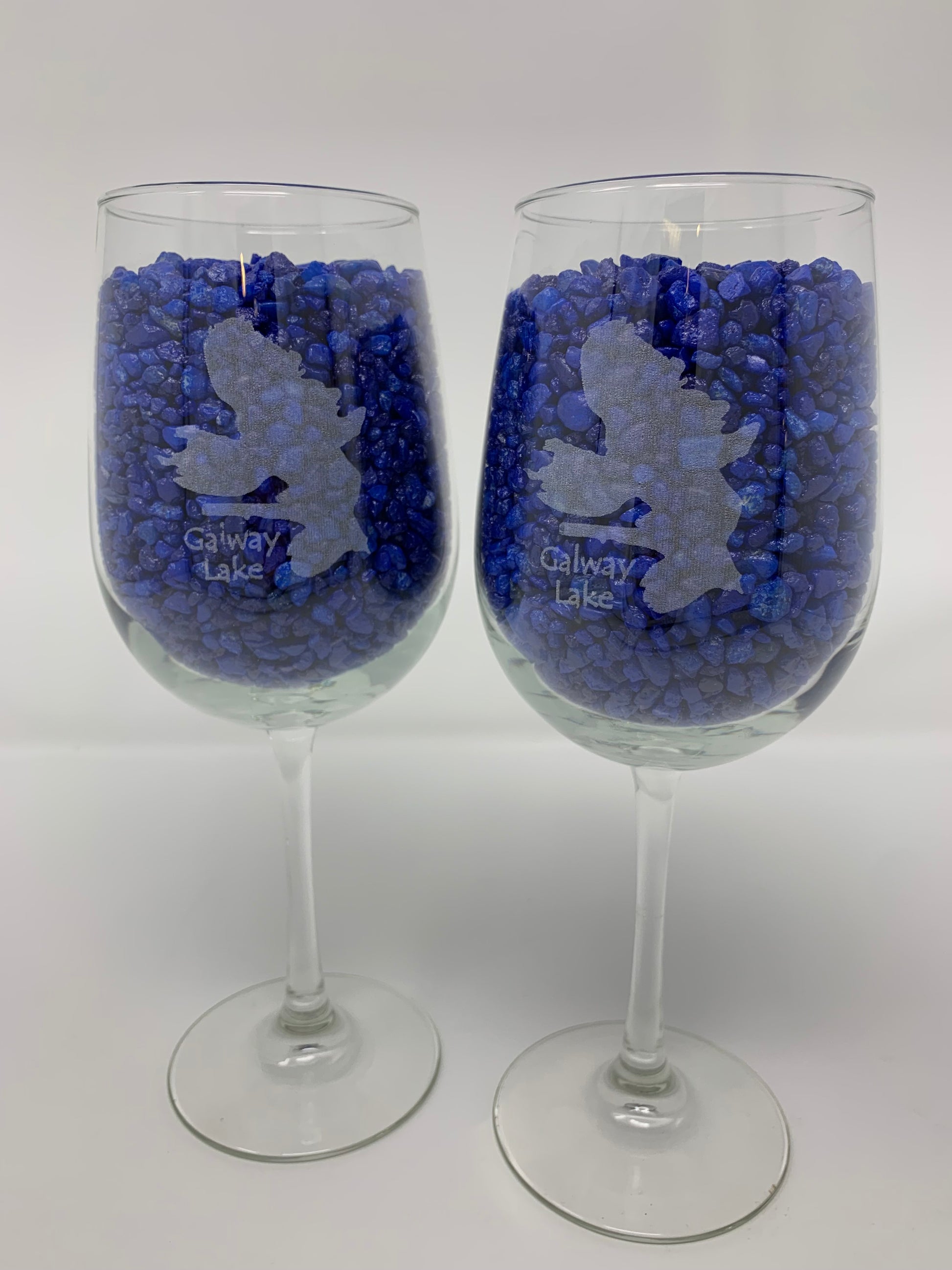 The Time Square Clock Shop TS Wine Glasses - Custom Set of 2 features each wine glass etched with a leaping person and "Galway Lake," filled with small blue stones, shown against a plain white background.
