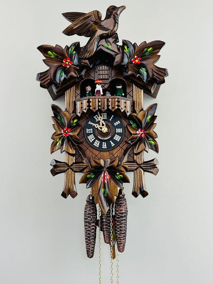The River City Cuckoo Clock - Dancers (MD411-14P) by River City Clocks features hand-carved leaf and bird details, colorful accents, pinecone weights, a black face with gold numbers, and small figurines above the clock.