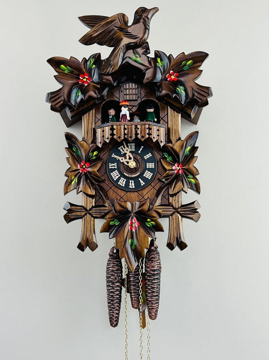 The River City Cuckoo Clock - Dancers (MD411-14P) by River City Clocks features hand-carved leaf and bird details, colorful accents, pinecone weights, a black face with gold numbers, and small figurines above the clock.