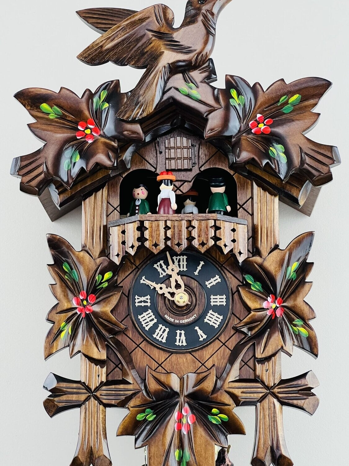 The River City Clocks MD411-14P features hand-carved leaves, a bird on top, and three figurines on a balcony above the dial showing 9:48; red and green accents complete this charming German cuckoo clock.