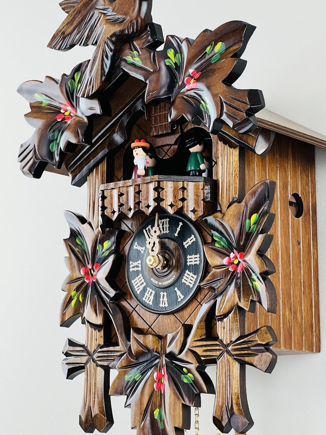 The River City Cuckoo Clock - Dancers (MD411-14P) by River City Clocks features hand-carved leaves, hand-painted red flowers, and two figurines on a balcony above a Roman numeral clock face with ornate hands.
