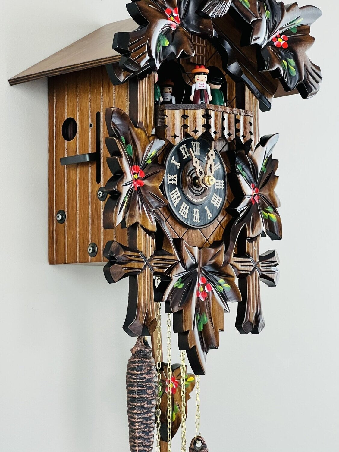 The River City Cuckoo Clock - Dancers (MD411-14P) by River City Clocks features carved leaves, red accents, a figure and bird above the face, and hanging chains and weights. This decorative clock mounts on your wall.