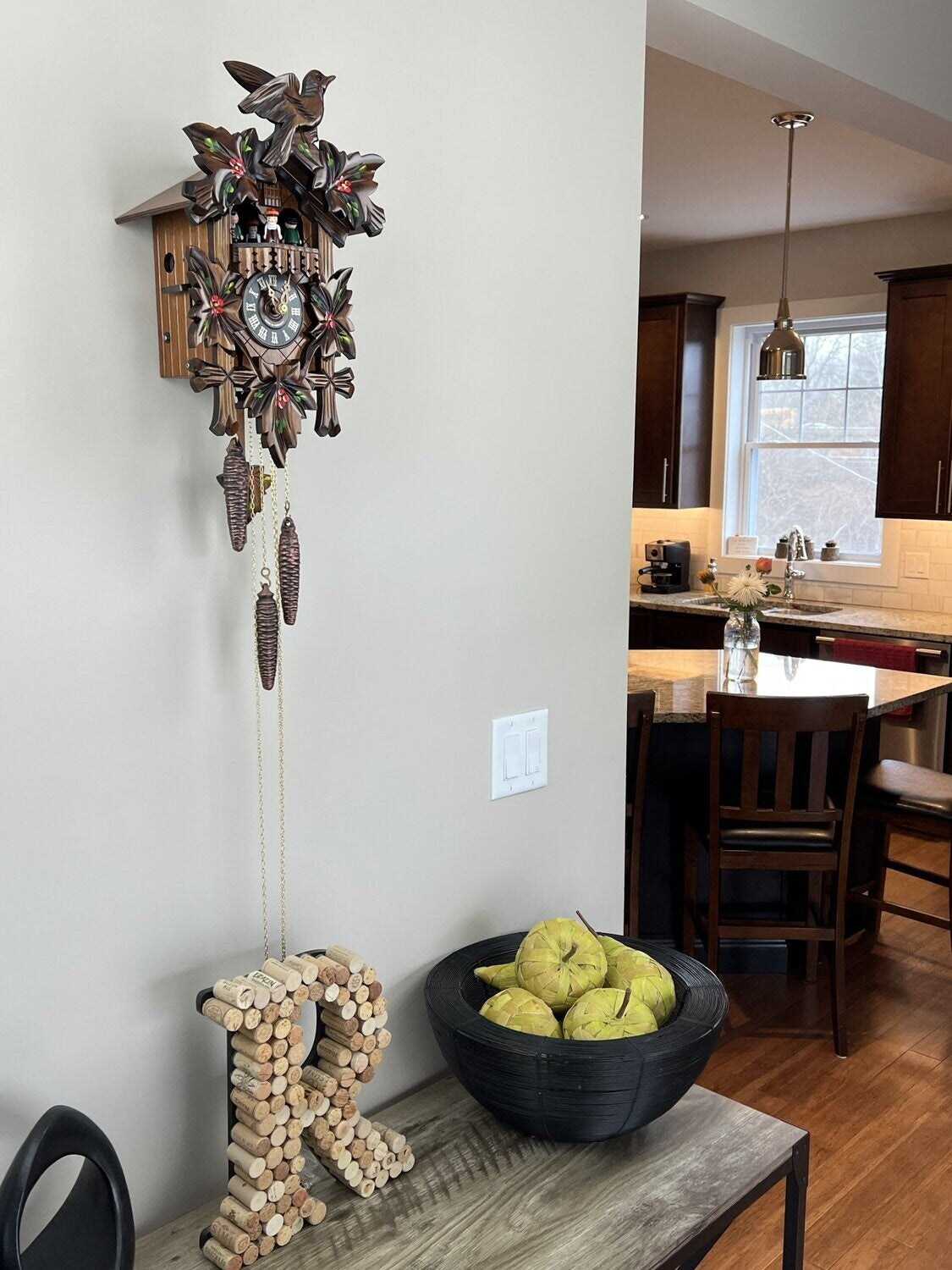 A River City Clocks River City Cuckoo Clock - Dancers (MD411-14P) with carved leaves and bird hangs above a black bowl of yellow pears and a wine cork letter R, with a kitchen featuring dark cabinets in the background.