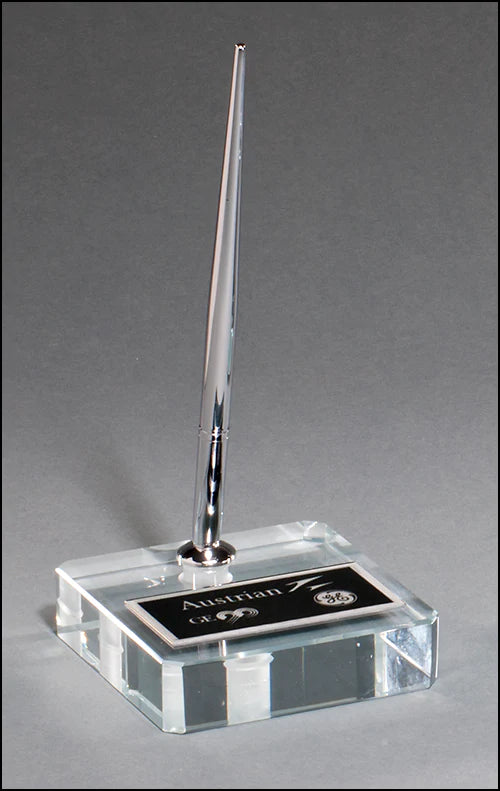 The Time Square Clock Shop Pen Set - Glass Base (PS8200) features a sleek silver pen standing upright in a clear, laser-engraveable base with a black plaque for text and logos, shown on a plain gray background.
