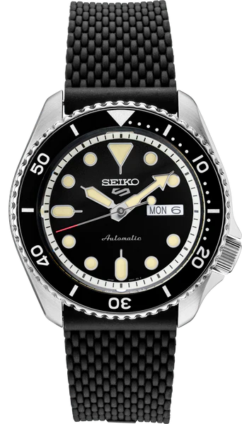 The Seiko Watch - SRPD95 - Seiko 5 Sports by Seiko showcases a round black sunray dial with luminescent markers, a day-date window at the 3 o’clock position, and is housed in a stainless steel case paired with a textured black silicone strap, displayed against a plain light gray background.