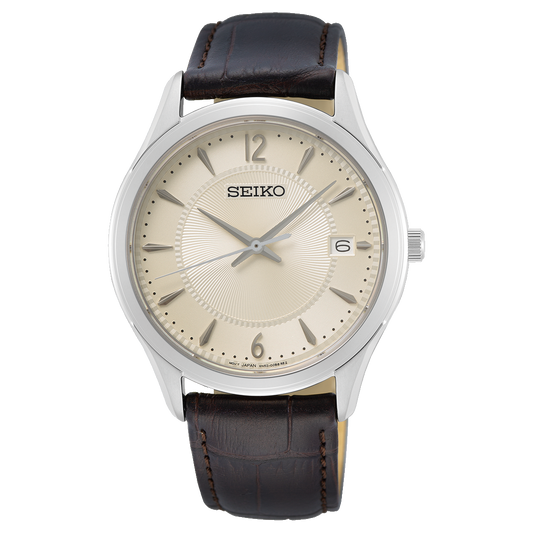 The Seiko Watch - Men's Essentials - SUR421 features a silver case, cream dial with numbers and markers, silver hands, date display at 3 o'clock, and a brown leather strap.