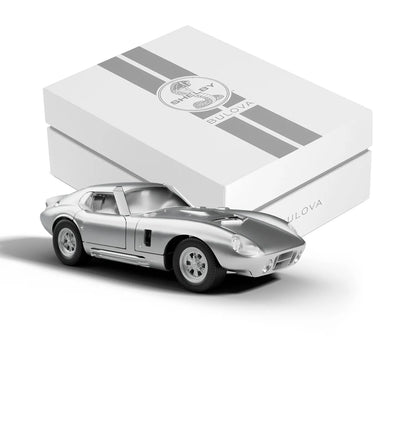 A silver die-cast toy sports car in front of a white Bulova SHELBY® Racer Chronograph - 96K118 presentation box with two grey racing stripes.