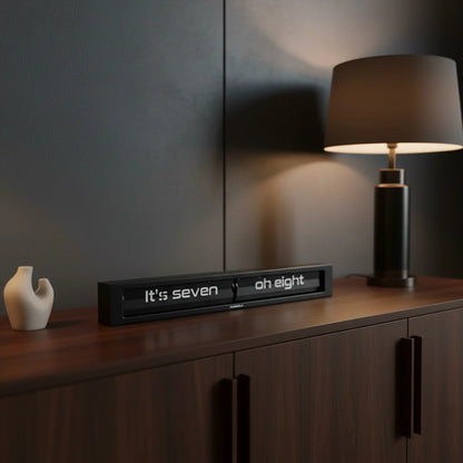 A modern lamp and a small white sculpture sit on a dark wood cabinet. On the cabinet, the Cloudnola Texttime Black Flip Clock (model 0072) displays the time as seven oh eight. The background features a dark, matte wall.