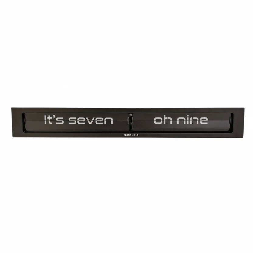 The Cloudnola Texttime Black Flip Clock (model 0072) by Cloudnola is a rectangular black metal wall clock that displays the time in words, such as “Its seven oh nine.” White text appears on a dark background within the clock’s frame, giving this modern flip clock a sleek and contemporary aesthetic.