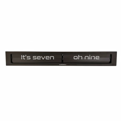 The Cloudnola Texttime Black Flip Clock (model 0072) by Cloudnola is a rectangular black metal wall clock that displays the time in words, such as “Its seven oh nine.” White text appears on a dark background within the clock’s frame, giving this modern flip clock a sleek and contemporary aesthetic.