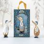 Two Enesco DCUK Ducks - Alpine Dinky Duck - 6017132 stand beside a festive gift box with a yellow ribbon handle and window, revealing the Alpine Dinky Duck ornament inside. A delightful wooden duck gift.