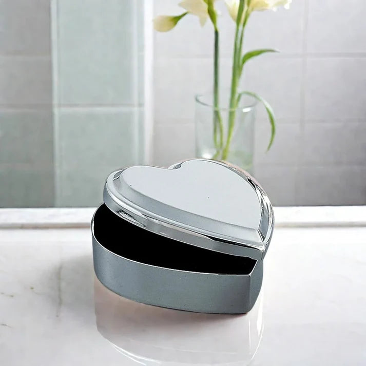 A Creative Gifts HEART JEWELRY BOX (22003) with a silver heart-shaped design and its lid slightly open rests on a marble surface, while a glass vase filled with white flowers appears softly blurred in the background.
