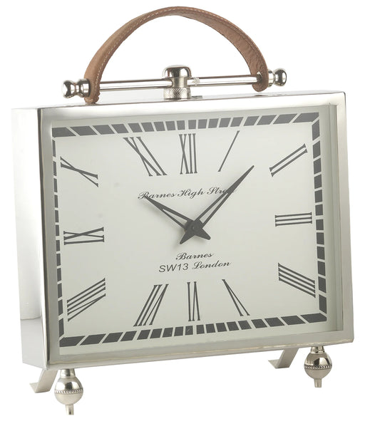 The A Sanoma - Nickel Clock - KG1003 features a rectangular nickel frame, Roman numerals, black hands, a decorative brown leather handle on top, and two small legs at the bottom, displaying 1:07. Brand: A Sanoma.