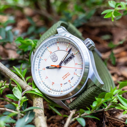 A Luminox Atacama Field Automatic XL.1907.NF silver watch with a white dial, orange second hand, green fabric strap, and day-date display is placed outdoors among green plants and brown mulch.