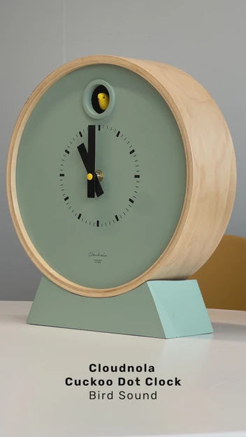 A modern round cuckoo clock by Cloudnola features a light wood frame and a sage green face, resting on a matching green base. A yellow and black cuckoo bird appears at the top. Below, the text reads: Cloudnola Clock – Sage Green Dot Cuckoo Clock – 0261.