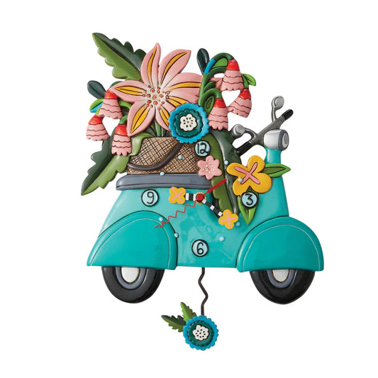 The Enesco Allen Designs Clock - Scoot-In Through Life (6012909) features a whimsical turquoise scooter with colorful flowers, leaves, and a straw hat, plus a clock face on the front wheel and a swinging flower pendulum below.