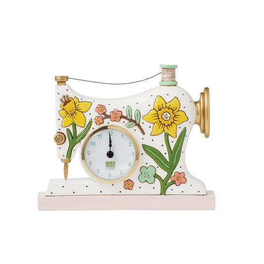The Enesco Allen Designs Clock – Sew Happy Desk Clock (6015369) features a whimsical vintage sewing machine design with white finish, yellow and pink flowers, green leaves, and gold accents. The clock face is cleverly set where the needle plate would be.